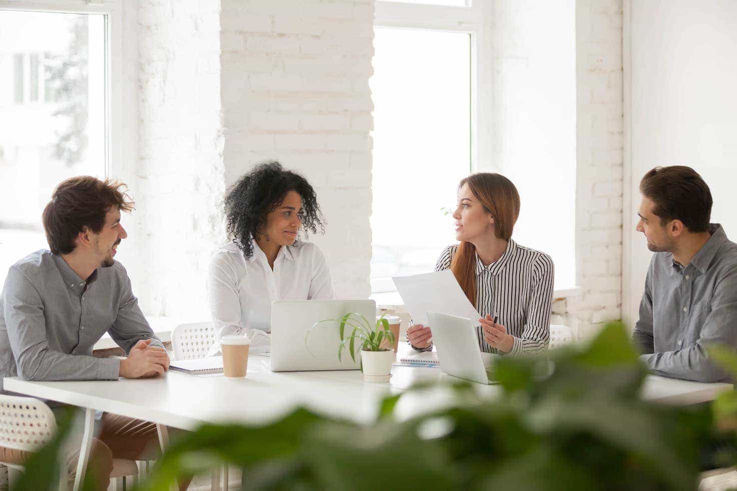 Quatre personnes sont assises autour d'une table en train de discuter dans une salle lumineuse. Une femme tient des feuilles de papier et s'adresse au groupe. Un ordinateur portable et des tasses à café sont disposés sur la table. - Article de blog Eventdrive