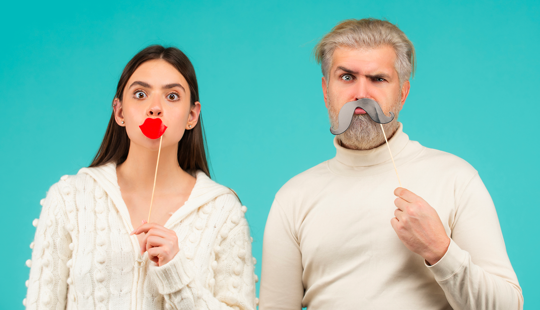 A young woman and an older man are playfully posing in front of a blue background. The woman is holding a red lip-shaped prop in front of her mouth, while the man is holding a fake gray mustache in front of his. Both have exaggerated facial expressions, with wide-open eyes, adding a fun and humorous touch to the image. They are wearing light-colored clothing, creating a contrast with the background. - Eventdrive Blog Article