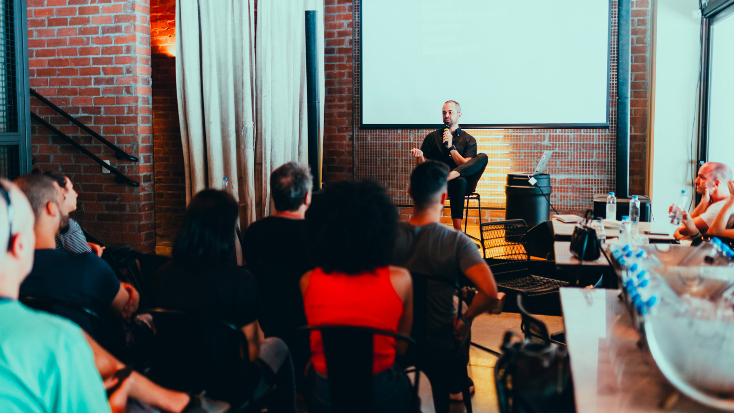 Conférencier assis sur une chaise devant un groupe de personnes lors d'un événement dans une salle en briques, avec un projecteur et des bouteilles d'eau visibles. - Article de blog Eventdrive