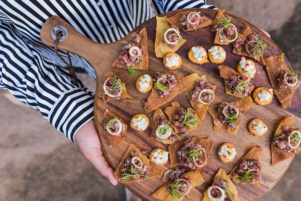 Une personne portant une chemise à rayures tient une planche en bois garnie de petits amuse-bouches. - Article de blog Eventdrive