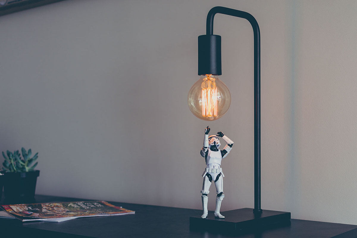 Une figurine de soldat en armure blanche est placée sous une lampe de bureau moderne avec une ampoule Edison exposée. La figurine semble lever les bras vers la lumière, créant une scène amusante. - Article de blog Eventdrive