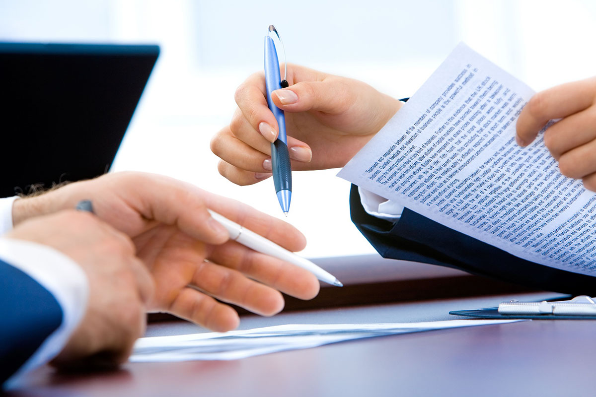 A Close-Up Shows Two People Discussing a Printed Document. One Is Holding a Sheet Filled With Text and a Blue Pen, While the Other Reaches Out With a White Pen. - Eventdrive Blog Article