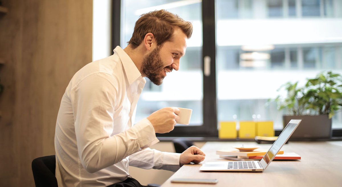 A smiling man, sitting at a desk, holds a cup of coffee while looking at his laptop screen. The scene takes place in a bright office with a green plant in the background, creating a professional yet relaxed atmosphere. - Eventdrive Blog Article