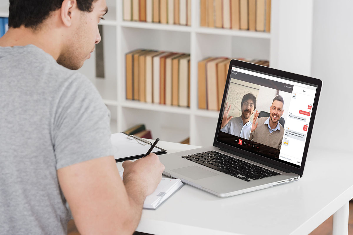 A man is participating in a videoconference on his laptop while taking notes on a notepad. Two other participants appear on the screen, smiling and waving. - Eventdrive Blog Article