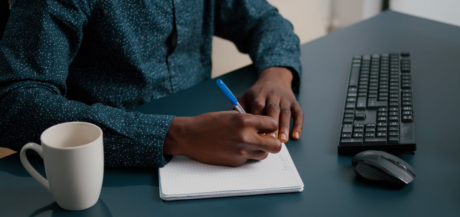 A person in a patterned shirt is writing notes in a notebook with a blue pen, seated at a desk. - Eventdrive Blog Article