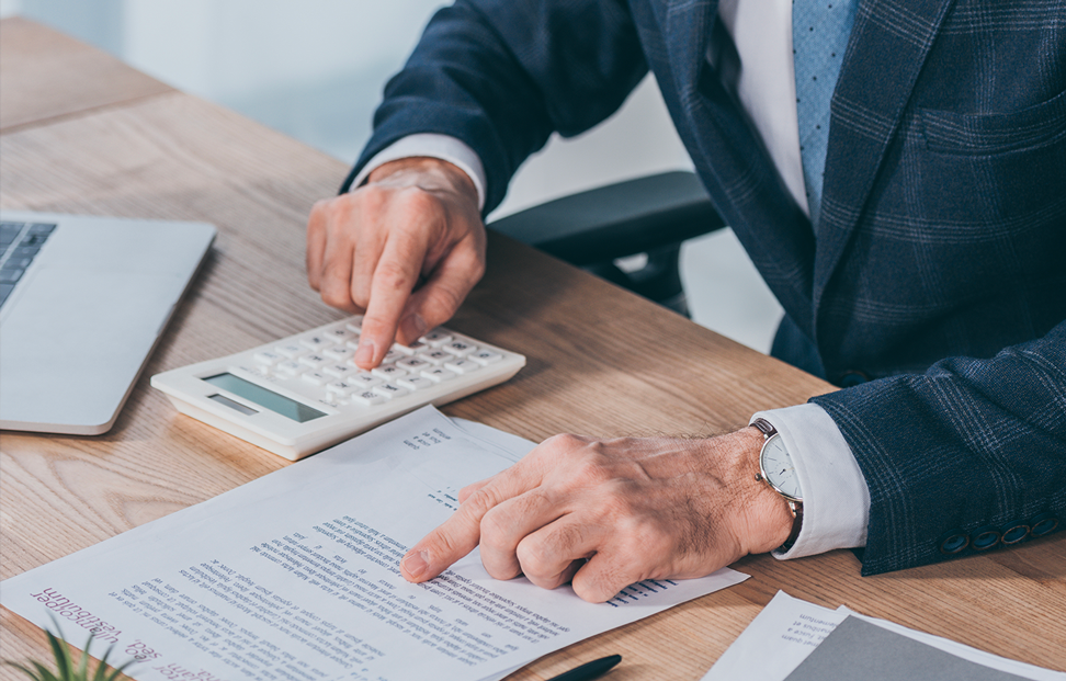 A man in a suit using a calculator and reviewing financial documents at his desk. He points to a line on the document while making calculations. A laptop is visible next to him on a wooden table. - Eventdrive blog article