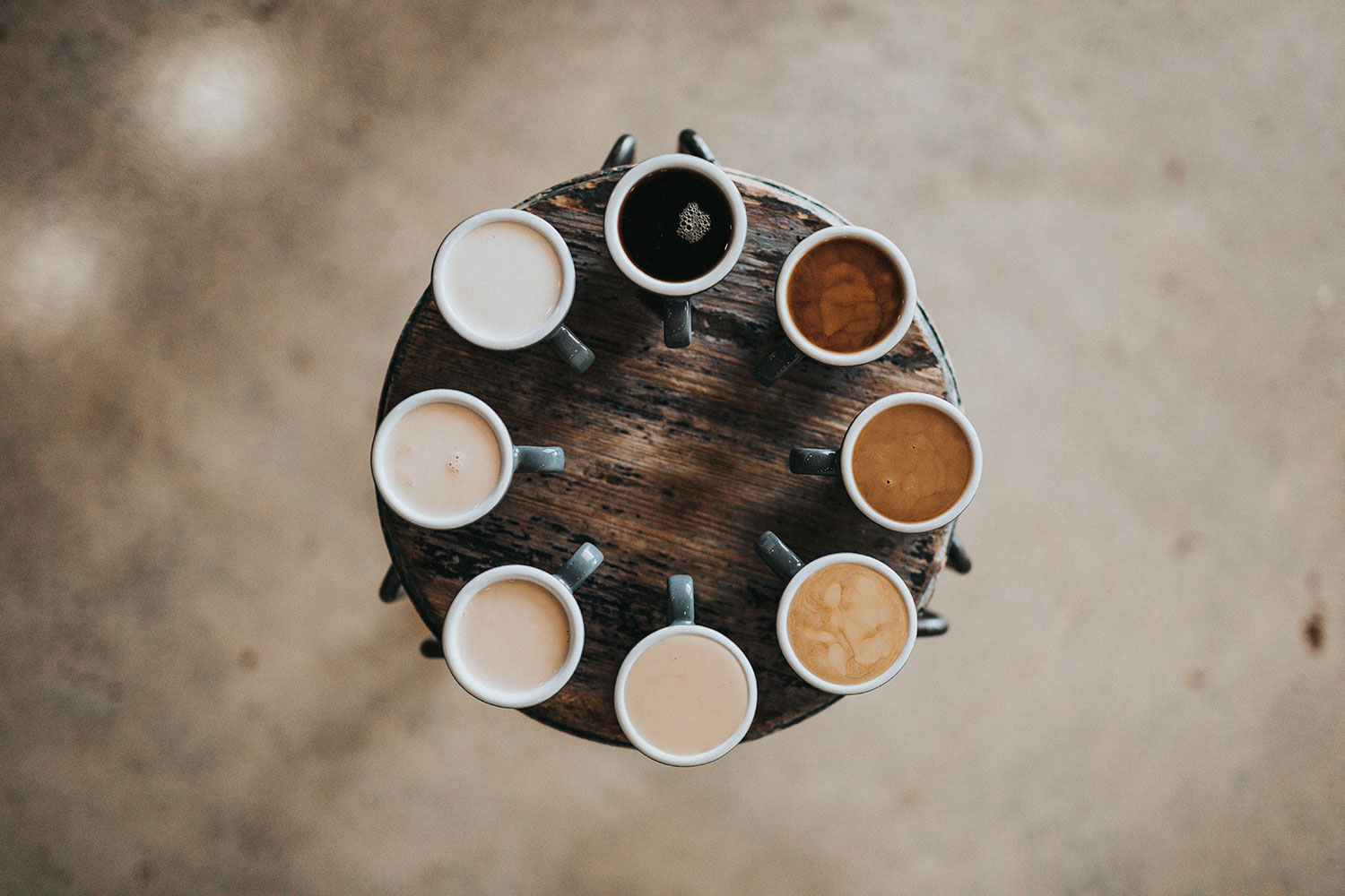 Vue aérienne de huit tasses de café disposées en cercle sur une table en bois. Les tasses contiennent différentes nuances de café, allant du plus clair au plus foncé, représentant une variété de préparations avec plus ou moins de lait. Le bois rustique de la table et la diversité des couleurs des cafés créent une composition visuelle harmonieuse et naturelle. - Article de blog Eventdrive