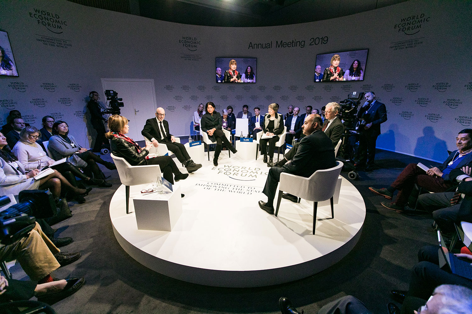 A group of participants sits in a circle during a session at the 2019 World Economic Forum, with several speakers discussing in the center of the room. Screens on the walls display the speakers, and cameras are recording the session. - Eventdrive Blog Article