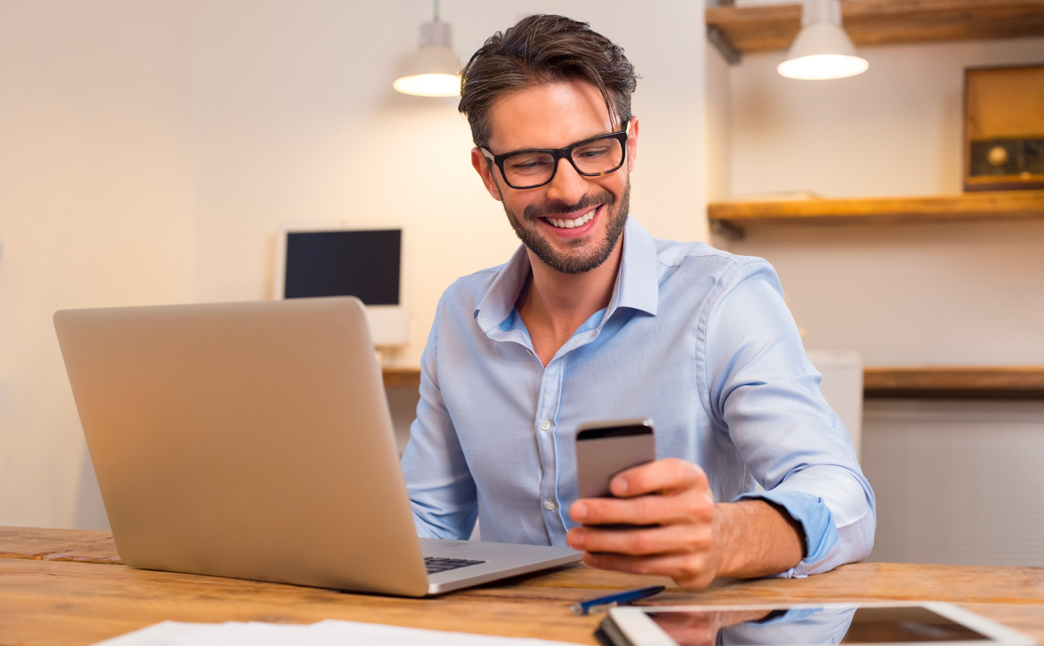 A man sitting at his desk, in front of his computer, smiles while attentively looking at his mobile phone.