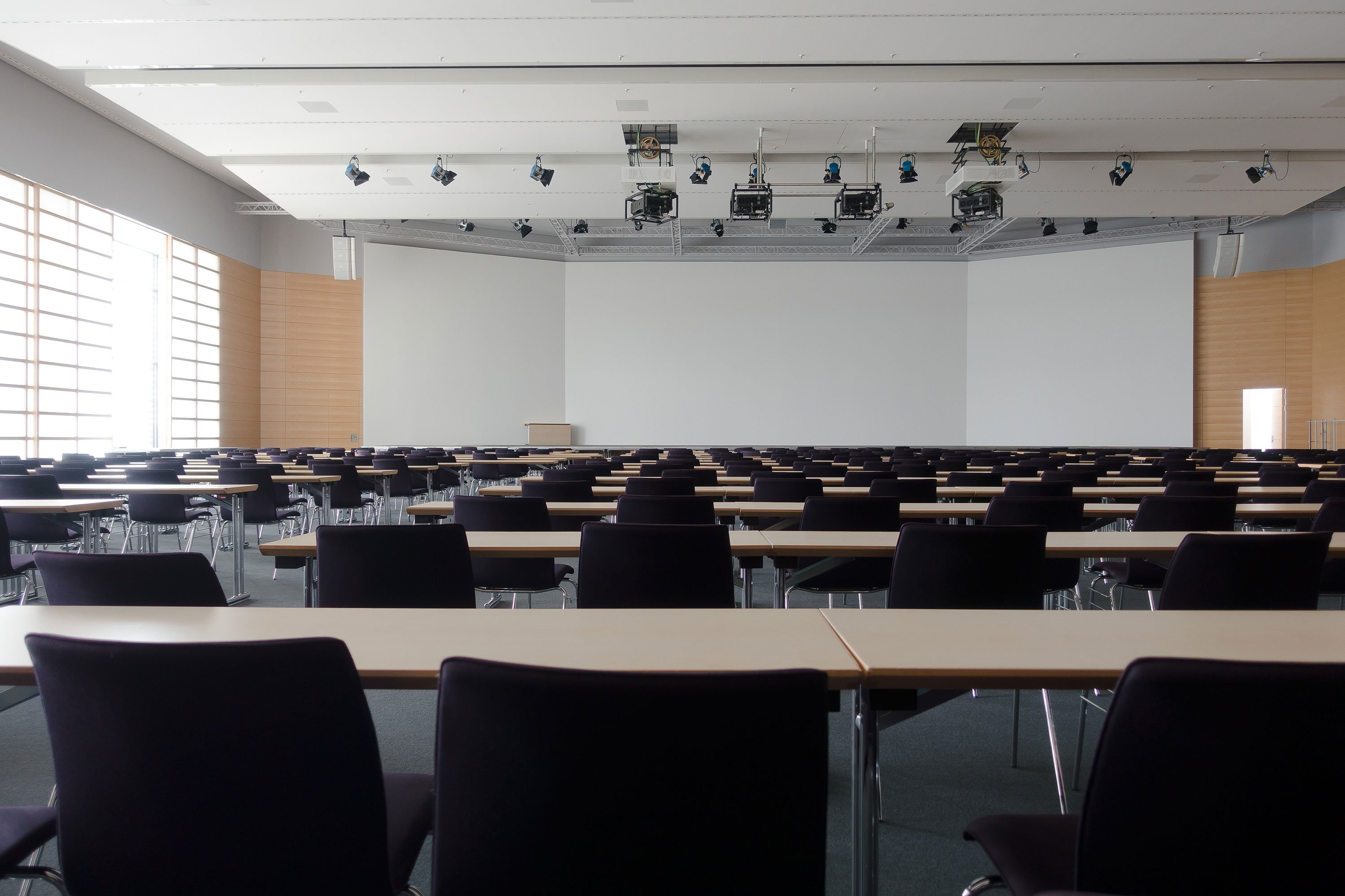 Spacious and modern conference room with rows of empty tables and chairs, equipped with projectors suspended from the ceiling.