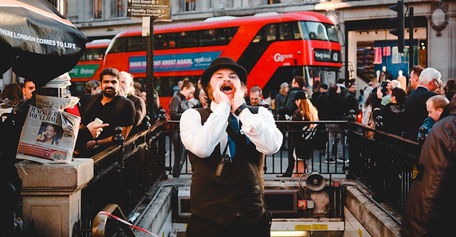 Un homme vêtu d'un chapeau et d'une cravate crie dans la rue, probablement pour attirer l'attention, devant une station de métro à Londres. En arrière-plan, un bus rouge à deux étages passe, entouré de passants et de touristes. - Article de blog Eventdrive