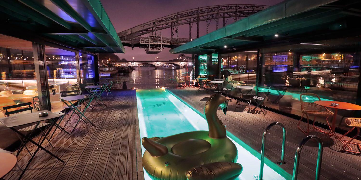 Le Restaurant Off Paris Seine, un bar flottant sur la Seine, propose un cadre moderne avec une piscine centrale, entourée de murs vitrés et d'un toit rétractable. Un grand cygne gonflable flotte dans la piscine, avec des sièges confortables de chaque côté, et une vue sur la rivière et un pont au loin. L'ambiance est chic et décontractée. - Article de blog Eventdrive