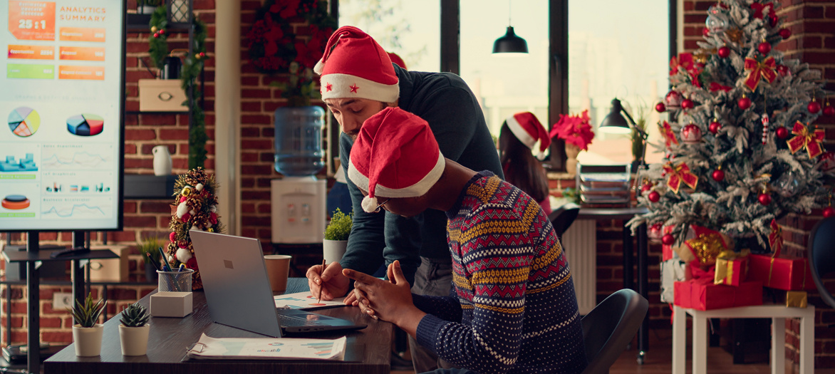 Deux hommes portant un pull et un bonnet de père Noël sont devant un bureau sur lequel se trouve un ordinateur.