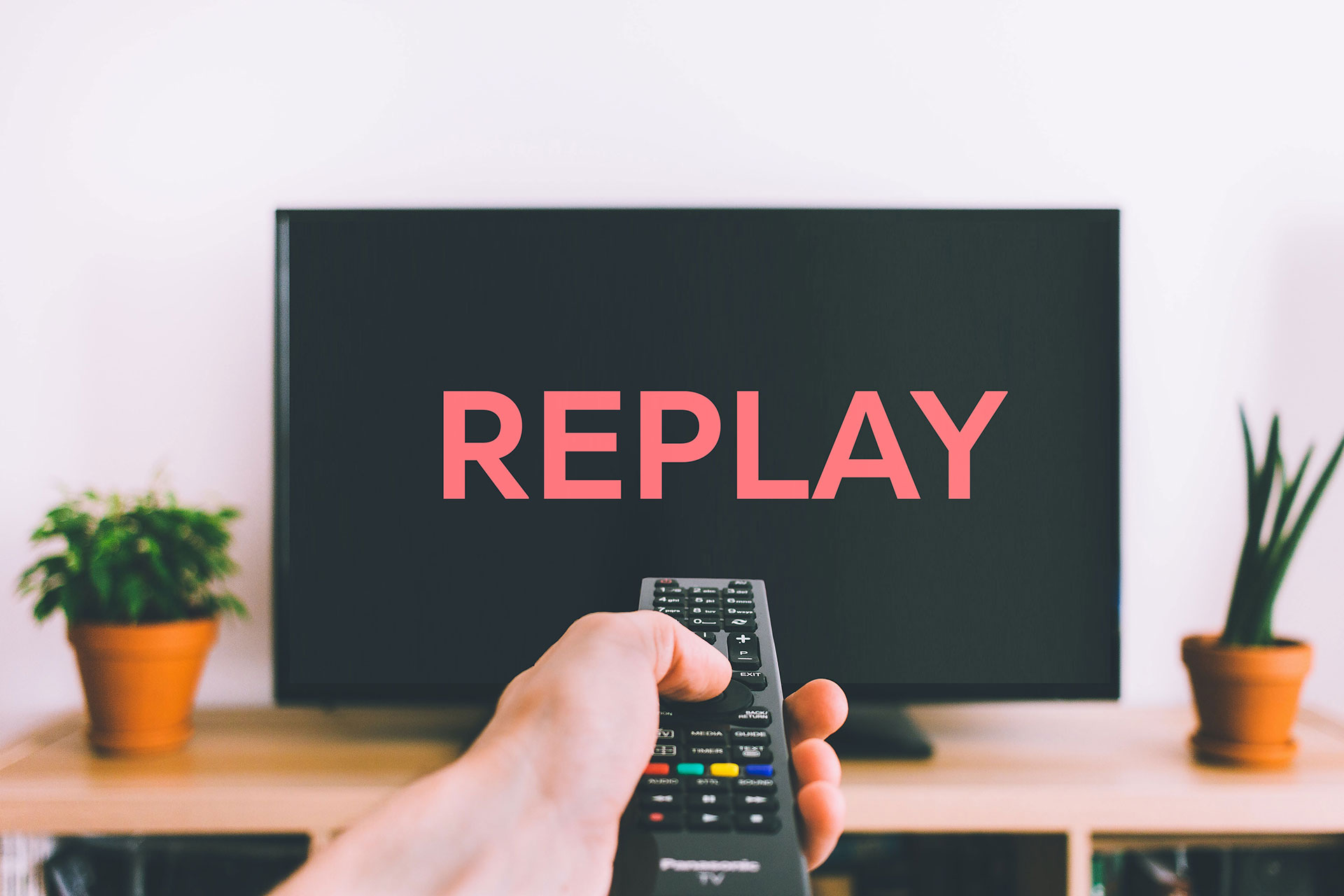 A man is holding a remote control. In front of him, the TV displays the word 