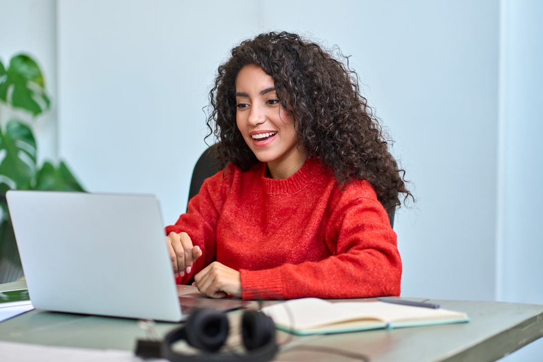 A Smiling Woman With Curly Hair Is Working on a Laptop, Sitting at a Desk With Headphones, a Notebook, and a Pen Nearby. - Eventdrive Blog Article