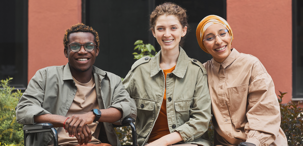 Trois personnes souriantes assises côte à côte en plein air. Un homme en fauteuil roulant, portant des lunettes et une veste verte, est assis à côté de deux femmes. - Article de blog Eventdrive