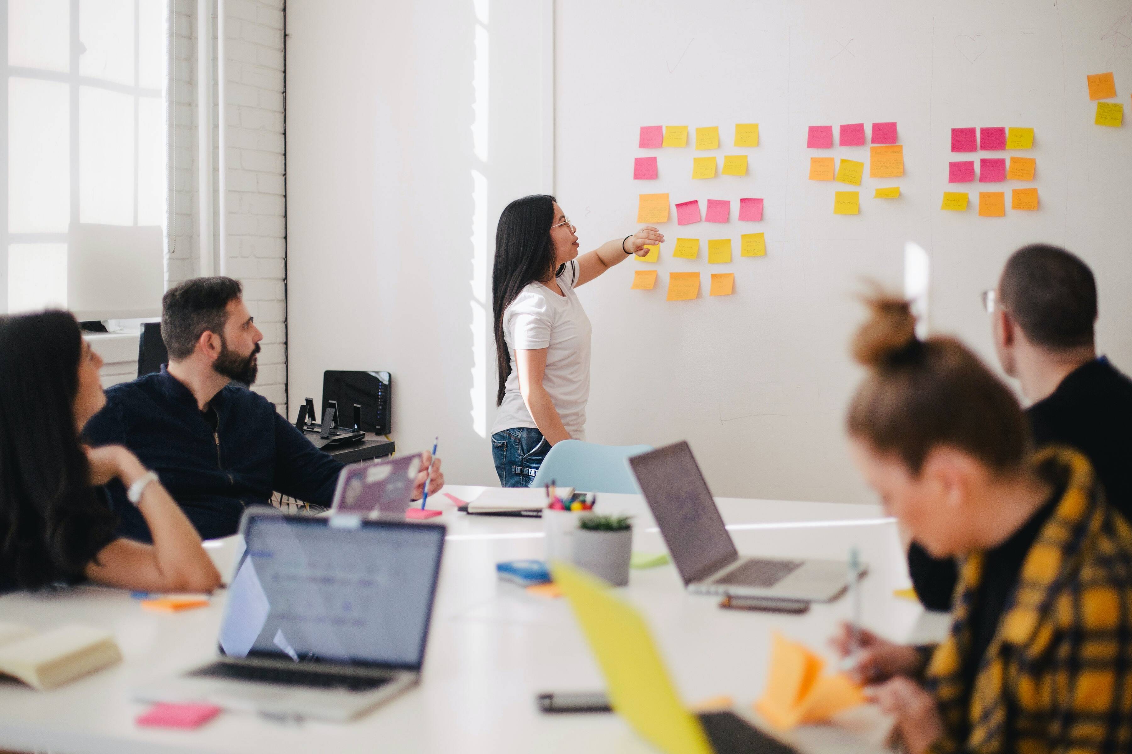 A group of people is gathered in a meeting room, where a woman is presenting ideas using colorful post-it notes stuck on a whiteboard. - Eventdrive Blog Article