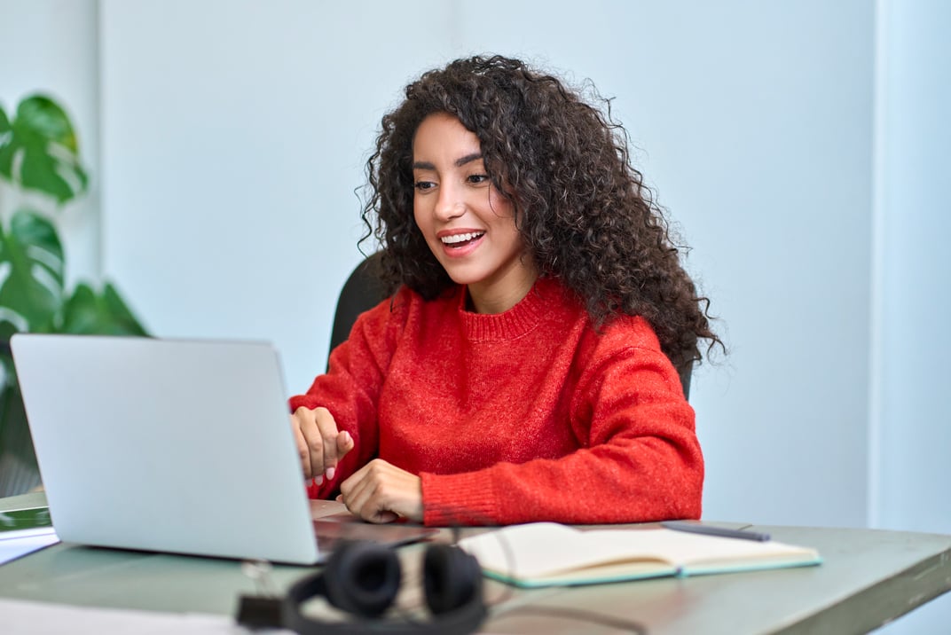 Une femme souriante aux cheveux bouclés travaille sur un ordinateur portable, assise à un bureau avec des écouteurs, un carnet et un stylo à proximité. - Article de blog Eventdrive